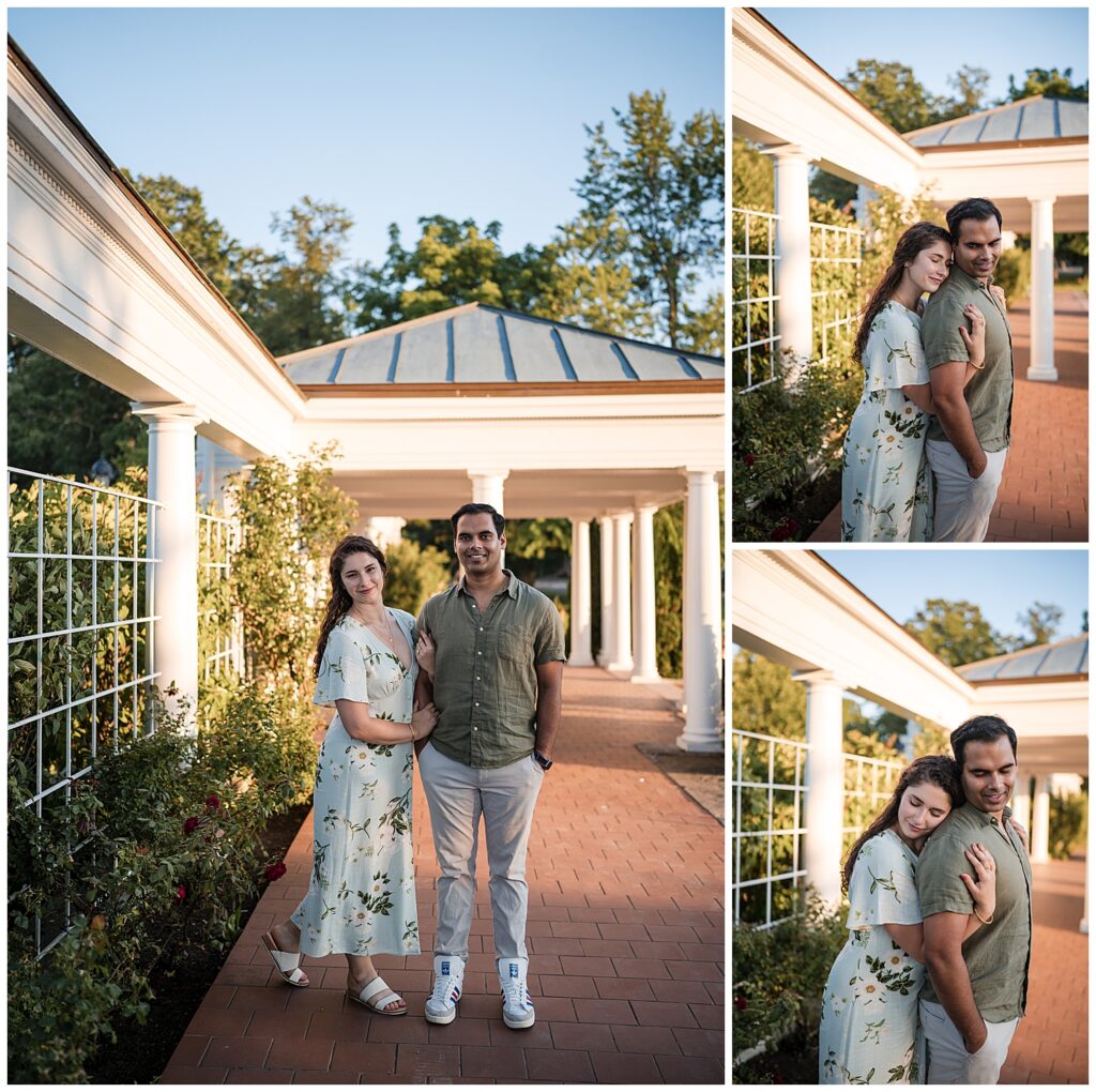 Couple stands leaning on each other in the Delaware Park rose Garden pavilion. Sarah is wearing a floral dress while Ankur is in a green linen button up shirt and gray pants.
