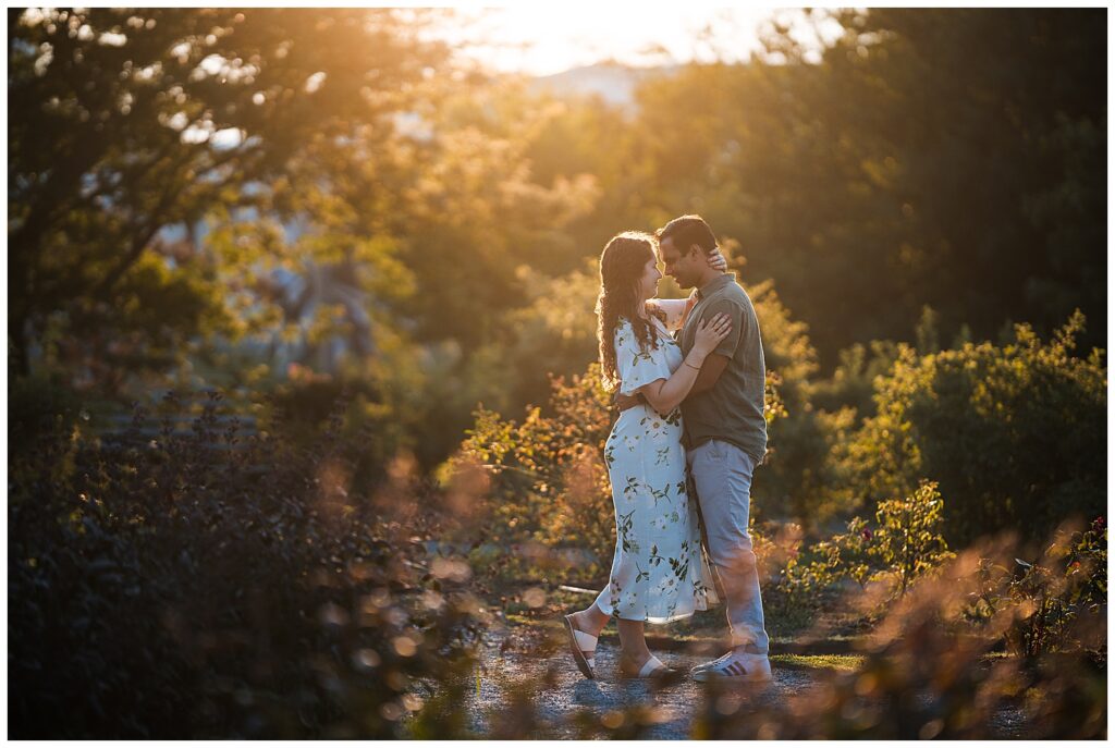 Sarah and Ankur stand facing each other in the Delaware Park Rose garden during sunset. Golden light surrounds them.