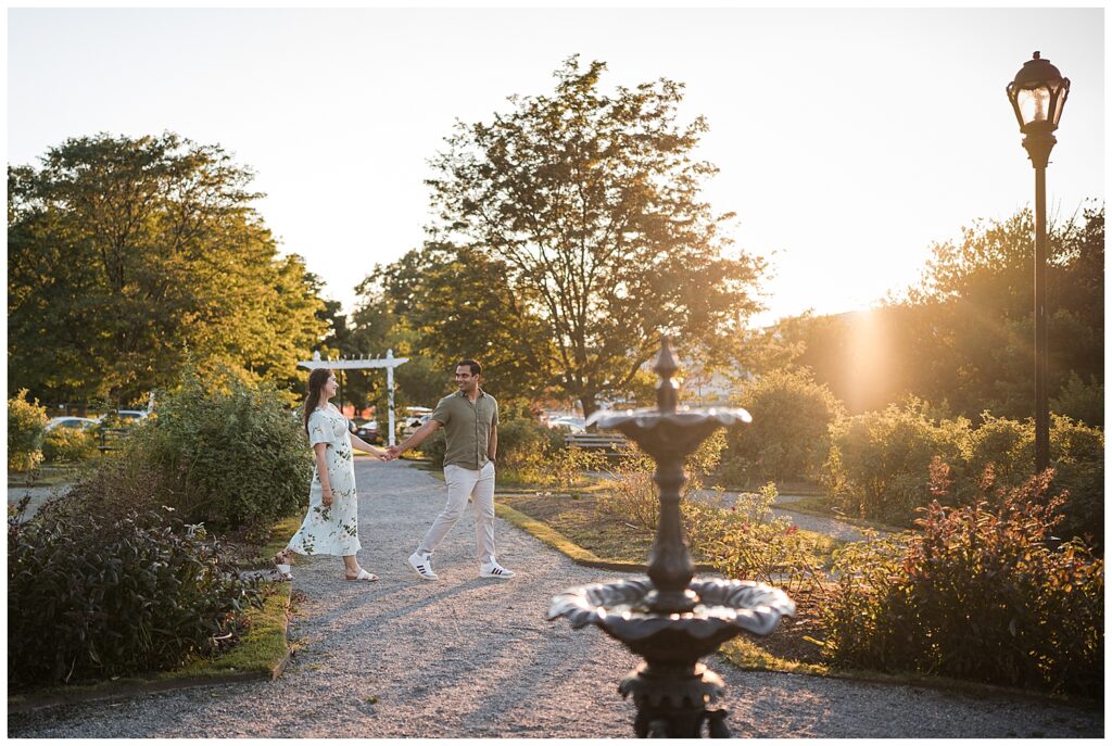 Ankur leads Sarah walking past the fountain in the Delaware Park rose garden during sunset.