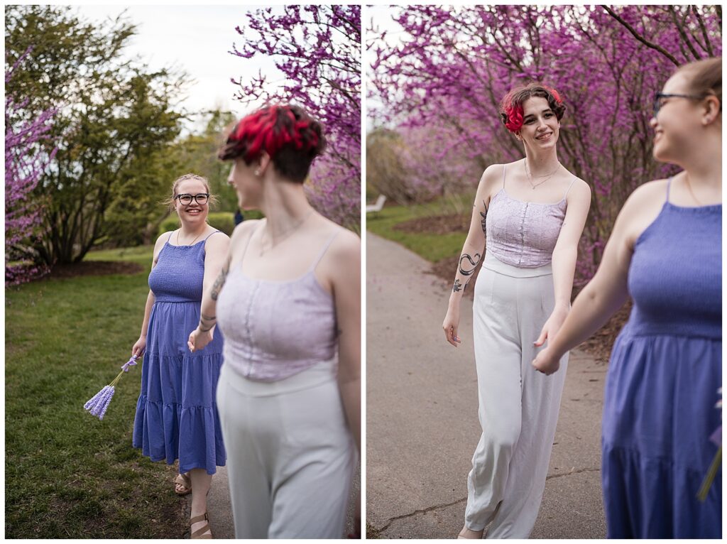 Cas and Kaylie lead each other walking forward down a path of highland park. Pink lilacs bloom in the background.