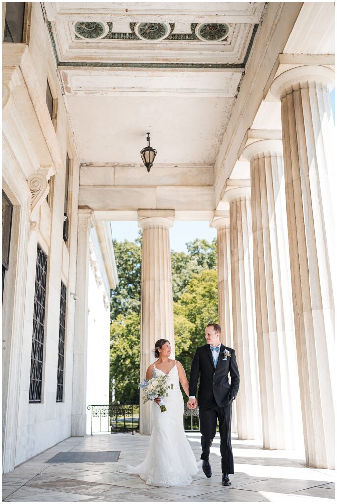 bride and and groom pose for formal photos on wedding day outside the buffalo history museum. They walk along the columns on the back of the building