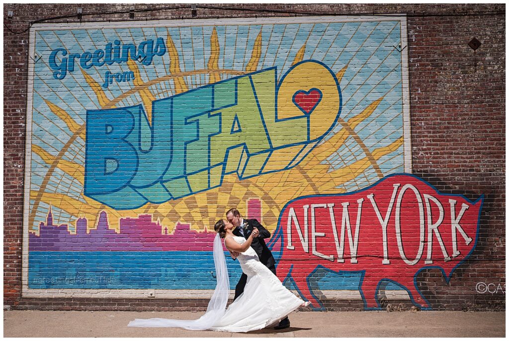 bride and groom dip for a kiss in front of the greetings from buffalo mural